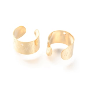 May include: Two gold-colored ear cuffs. Each cuff is a curved band with a star pattern. The cuffs are open at one end and have a smooth, shiny finish. One cuff has a small hole.