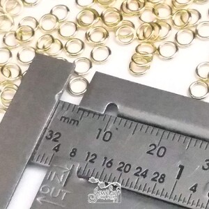 K&ouml;nnte beinhalten: Eine Ansammlung kleiner, kreisf&ouml;rmiger, goldfarbener Metall-Biegeringe, die auf einer Fl&auml;che verstreut sind. Ein Metall-Messschieber ist &uuml;ber den Ringen positioniert und zeigt ein Ma&szlig; von etwa 4 Millimetern auf der metrischen Skala.