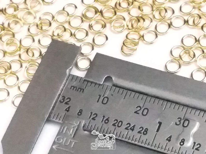 K&ouml;nnte beinhalten: Eine Ansammlung kleiner, kreisf&ouml;rmiger, goldfarbener Metall-Biegeringe, die auf einer Fl&auml;che verstreut sind. Ein Metall-Messschieber ist &uuml;ber den Ringen positioniert und zeigt ein Ma&szlig; von etwa 4 Millimetern auf der metrischen Skala.