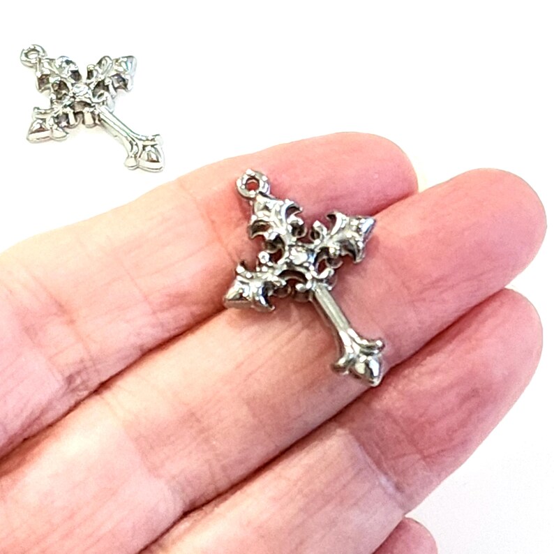 May include: Ornate silver-colored cross pendant with detailed floral and scrollwork design. The cross has a loop at the top for attaching to a chain or cord. The pendant is held in a hand.