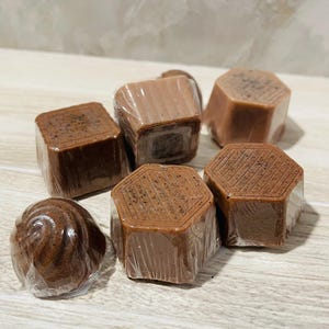 May include: An assortment of individually wrapped, dark brown, chocolate-colored soaps. The soaps have various shapes, including square, hexagonal, and a spiral design. The soaps are packaged in clear plastic wrap.