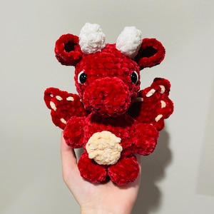 May include: A handmade red crochet dragon with white horns and wings. The dragon has black eyes and a tan belly. The wings have white stitching. This plush toy is a cute and cuddly creature.