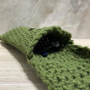 May include: A green crocheted pouch, open to reveal a pair of black glasses. The pouch is made of a textured, olive-green yarn and has a small metal clasp. The glasses are partially visible inside.