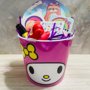 May include: A pink gift bucket with a cartoon face and yellow bow detail, filled with a red rubber duck, stickers, and a face mask. The bucket has the words "Kid Size Mask!" on the mask packaging.