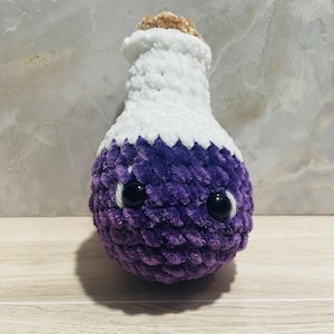 May include: A plush, handmade potion bottle in purple and white. The bottle has a rounded base and a long neck, with a cork stopper. The bottle has two large, black, googly eyes, giving it a whimsical appearance. The plush is made of soft, textured yarn.