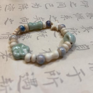 May include: A handmade bracelet featuring a mix of ceramic beads in white, light blue, and pale green. The bracelet includes star, flower, and bone-shaped beads, strung together with a neutral cord. The design is simple and elegant.