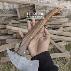 Könnte beinhalten: Eine handgefertigte Axt mit Holzgriff und einer Stahlklinge mit Muster. Der Griff ist hellbraun mit Holzmaserung. Die Klinge hat ein einzigartiges Design. Der Text "DELTAONE KNIVES & TOOLS" ist sichtbar.