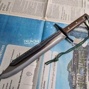 May include: A large, handmade knife with a dark wooden handle and a long, silver blade. The knife has a black crossguard and a green cord attached to the handle. The knife is resting on a newspaper with the text "DELTAONE KNIVES & TOOLS".