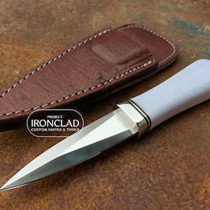 May include: A custom knife with a white handle and a polished steel blade, next to a brown leather sheath. The knife has a silver bolster and a sharp point. The sheath is stitched with white thread. The knife is labeled "PROJECT IRONCLAD CUSTOM KNIVES & TOOLS."