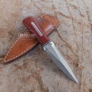 May include: A custom knife with a polished steel blade and a dark wood handle with a silver accent. The knife rests on a brown leather sheath with white stitching. The logo "IRONCLAD CUSTOM KNIVES & TOOLS" is visible.