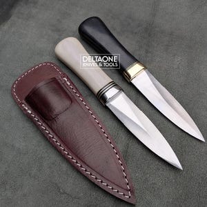 May include: Two fixed-blade knives with pointed steel blades and a brown leather sheath. One knife has a cream-coloured handle with silver-tone bolsters, while the other has a black handle with gold-tone bolsters. Text reads DELTAONE KNIVES & TOOLS.