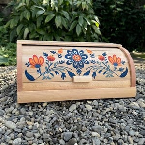 May include: Wooden bread box with a floral design in orange, blue, and white. The box has a curved top and a small wooden handle. The bread box is made of light-colored wood and sits on a bed of small gray stones.