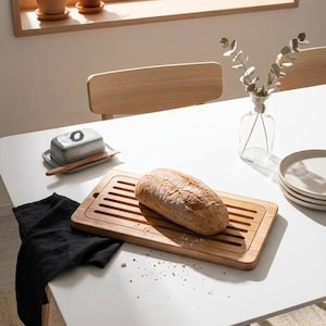 May include: A wooden breadboard with a loaf of bread sits on a white table, next to a butter dish and a black cloth. A vase with greenery and plates are also on the table. The scene is set in a bright room with natural light.