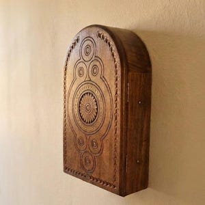 May include: A dark brown wooden wall-mounted cabinet with an arched top. The front panel features intricate carved circular patterns and a central flower motif. The cabinet has a hinged side door with visible metal hinges and a smooth wood grain finish.