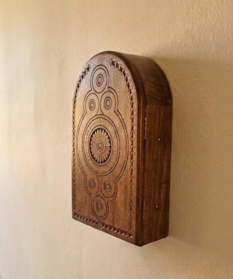 May include: A dark brown wooden wall-mounted cabinet with an arched top. The front panel features intricate carved circular patterns and a central flower motif. The cabinet has a hinged side door with visible metal hinges and a smooth wood grain finish.