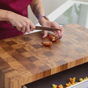 May include: A wooden butcher block cutting board with a pull-out tray. A person is slicing a red tomato on the board with a knife. Chopped vegetables are in the tray. The board is made of various shades of brown wood.