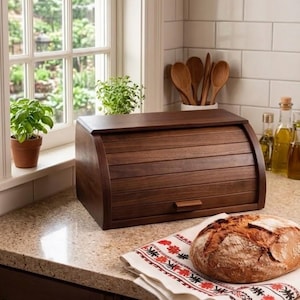 May include: A dark brown wooden bread bin with a roll-top lid is on a kitchen counter, next to a loaf of bread on a patterned cloth. A small potted plant is on the windowsill. Wooden spoons and olive oil bottles are also visible.