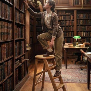 May include: A person wearing a brown cardigan, a patterned button-down shirt, and brown trousers stands on a wooden step stool. The person is reaching toward a tall wooden bookshelf filled with many books in a library setting with a green desk lamp.