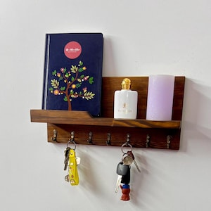 Può includere: Un portachiavi in legno marrone scuro con un ripiano, con un libro blu navy con un design ad albero floreale, una bottiglia di profumo bianca e una candela lavanda. Le chiavi sono appese ai ganci sottostanti.