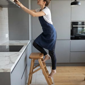 Può includere: Uno sgabello a gradini in legno marrone chiaro a tre livelli con un'apertura a maniglia sul gradino superiore. Lo sgabello presenta un design pieghevole con cerniere in metallo e una finitura in venature del legno naturale.