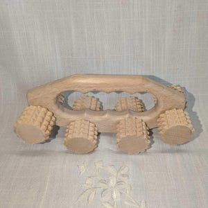 May include: A light-colored wooden handheld massage tool featuring a curved handle with a scalloped grip and six cylindrical rollers. Each roller has a textured, spiked surface designed for rolling across the skin to provide pressure.