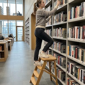 May include: A light-coloured wooden step stool with three tiers stands in a library aisle. The stool features a sturdy frame with angled legs and smooth, rounded steps, providing elevation to reach books on the high shelves of the tall bookcases.