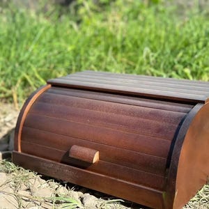 May include: A dark brown wooden bread bin featuring a roll-top lid with horizontal slats and a small rectangular handle centred on the front panel. The container has a curved side profile and a flat top surface, constructed from stained wood.