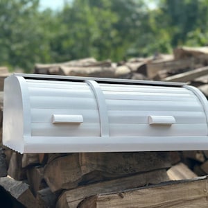 May include: A white, roll-top bread box with a curved top and two small handles. The box is made of wood and has a slatted design on the front. It sits on a pile of firewood, with a blurred green background.