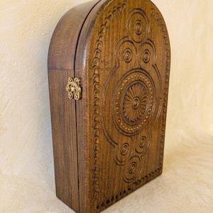 May include: A dark brown wooden box with an arched top, featuring intricate circular carvings and a central sunburst pattern on the front panel. The box is secured with a small, metallic brass-colored latch on the side, showcasing a smooth finish.