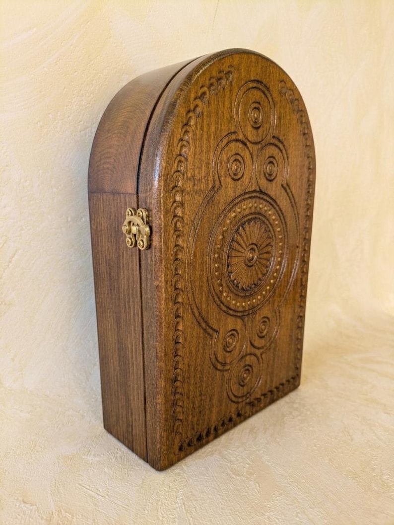 May include: A dark brown wooden box with an arched top, featuring intricate circular carvings and a central sunburst pattern on the front panel. The box is secured with a small, metallic brass-colored latch on the side, showcasing a smooth finish.