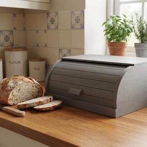 May include: A gray wooden bread box with a roll-top lid and a small handle, positioned next to a loaf of crusty bread, two sliced pieces of bread, and a metal bread knife with a wooden handle on a wooden kitchen countertop.