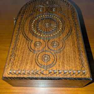 May include: A rectangular wooden box with a rounded top edge, featuring intricate dark brown carved geometric patterns and a central circular sunburst motif accented with small, evenly spaced brass-colored metal studs embedded in the wood.