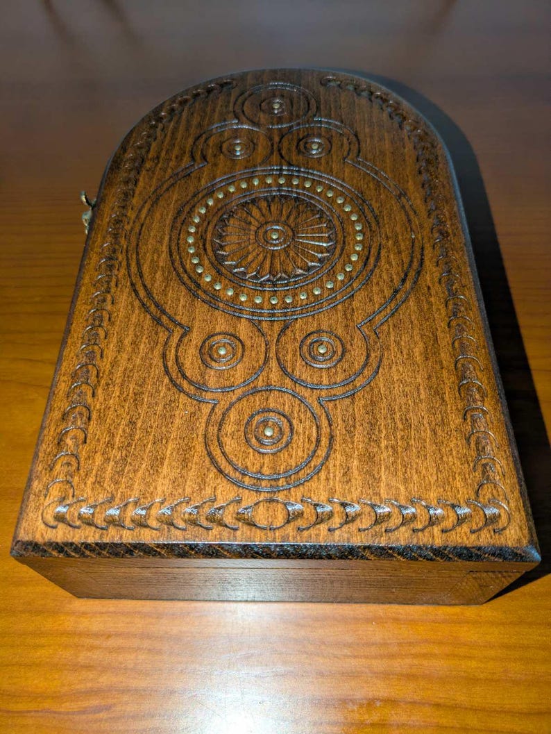 May include: A rectangular wooden box with a rounded top edge, featuring intricate dark brown carved geometric patterns and a central circular sunburst motif accented with small, evenly spaced brass-colored metal studs embedded in the wood.