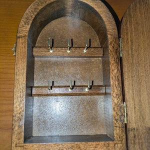 May include: A wall-mounted wooden key cabinet with an arched top, featuring a dark brown finish and a hinged door. The interior contains two horizontal wooden bars, each equipped with three small metal hooks for hanging keys.