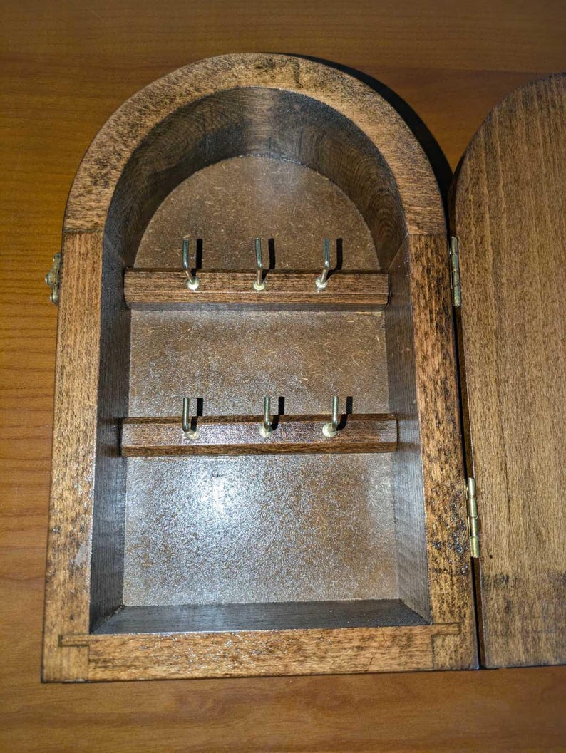 May include: A wall-mounted wooden key cabinet with an arched top, featuring a dark brown finish and a hinged door. The interior contains two horizontal wooden bars, each equipped with three small metal hooks for hanging keys.
