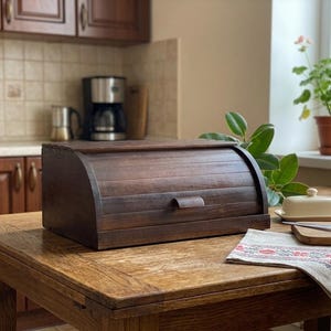 May include: A dark brown wooden bread bin with a roll-top lid and a small handle. The bin is on a wooden table. A glass of milk, a butter dish, and a decorative cloth are also on the table.