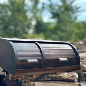 Op de afbeelding: Een donkerbruine houten brooddoos met een roll-top ontwerp. De doos heeft een gebogen bovenkant en een rechthoekige basis. De brooddoos staat op een stapel houten stammen. De achtergrond is wazig met groen gebladerte.