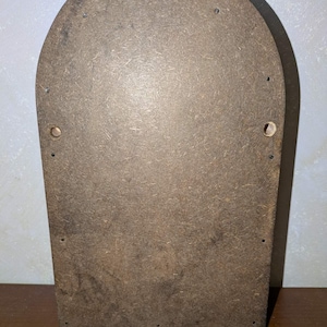May include: A brown, arched-top wooden board made of compressed particle material. The surface features a textured, speckled appearance with several small, evenly spaced metal nails along the perimeter and two circular holes on the sides.