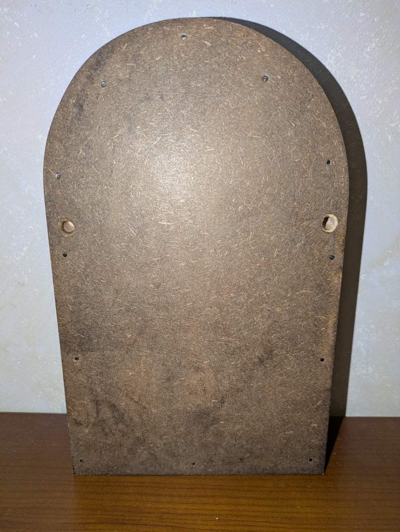 May include: A brown, arched-top wooden board made of compressed particle material. The surface features a textured, speckled appearance with several small, evenly spaced metal nails along the perimeter and two circular holes on the sides.
