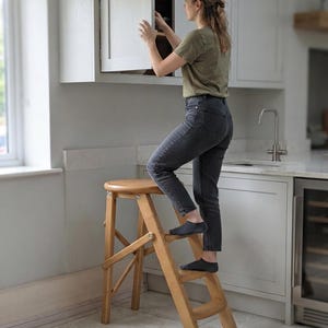 Puede incluir: Un taburete de madera de color marrón claro con un asiento redondo y dos peldaños inferiores. El taburete presenta un diseño plegable con soportes laterales articulados y patas inclinadas, proporcionando una plataforma estable para alcanzar zonas altas.