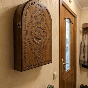 May include: A wall-mounted wooden cabinet with an arched top and a dark brown finish. The front panel features intricate carved geometric patterns, including a central flower motif surrounded by circular designs and small brass-colored studs.