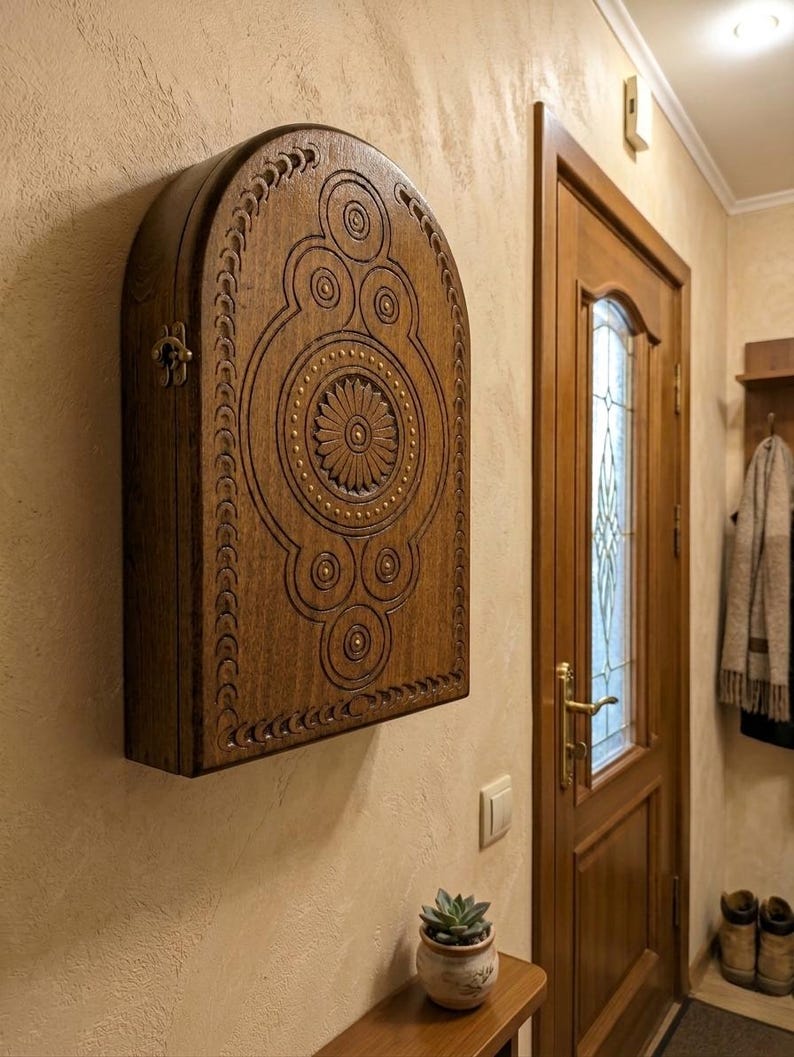 May include: A wall-mounted wooden cabinet with an arched top and a dark brown finish. The front panel features intricate carved geometric patterns, including a central flower motif surrounded by circular designs and small brass-colored studs.