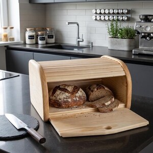 Peut inclure: Un coffre à pain en bois avec un couvercle roulant, ouvert pour révéler deux pains et plusieurs tranches. Le coffre est en bois clair et repose sur un comptoir sombre. Un couteau et une planche à découper sont au premier plan.