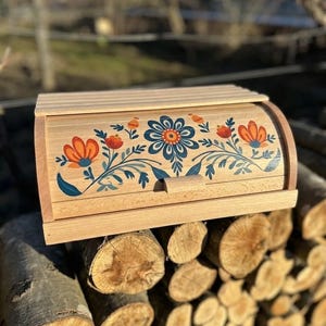 May include: A wooden bread box with a roll-top lid. The front of the box is decorated with a floral design in orange, blue, and teal. The box is made of light-colored wood and sits on a pile of cut logs.