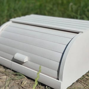 May include: A white wooden bread bin with a roll-top lid. The bin has a curved design and a small handle on the front. The bread bin is made of wood and painted white. The bin is sitting on a surface of dirt and grass.