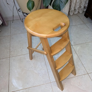 May include: A light-coloured wooden step stool with a round seat and a built-in handle. The stool has three steps and is designed to fold for storage. The overall height is approximately 61 cm.