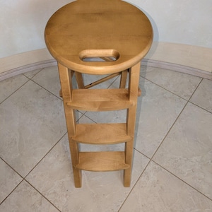Könnte beinhalten: Ein Holztritt mit rundem Sitz und drei Stufen. Der Hocker ist aus hellem Holz gefertigt und hat einen Griff, der aus dem Sitz geschnitten ist. Die Stufen sind gebogen und bieten Halt beim Klettern.