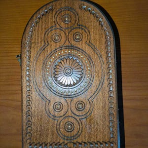 May include: A rectangular wooden box with an arched top, featuring a dark brown finish and intricate carved geometric patterns. The design includes a central floral motif surrounded by small brass-colored studs and a decorative border along the edges.