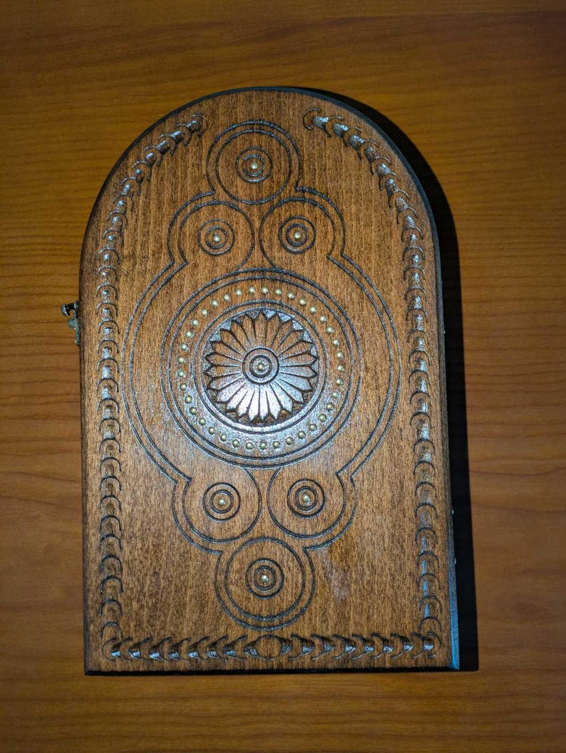 May include: A rectangular wooden box with an arched top, featuring a dark brown finish and intricate carved geometric patterns. The design includes a central floral motif surrounded by small brass-colored studs and a decorative border along the edges.