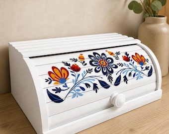 May include: White wooden bread box with a roll-top lid. The front is decorated with a floral design in blue, red, and yellow. The box has a white knob and is on a wooden surface.
